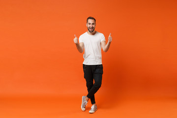Cheerful excited young man in casual white t-shirt posing isolated on bright orange wall background studio portrait. People sincere emotions lifestyle concept. Mock up copy space. Showing thumbs up. © ViDi Studio