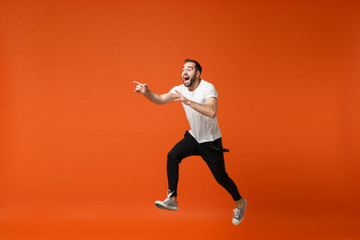 Fototapeta na wymiar Excited young man in casual white t-shirt posing isolated on orange wall background. People lifestyle concept. Mock up copy space. Having fun, fooling around, jumping, pointing index fingers aside.