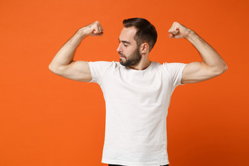 Strong attractive young man in casual white t-shirt posing isolated on orange wall background, studio portrait. People sincere emotions lifestyle concept. Mock up copy space. Showing biceps, muscles.