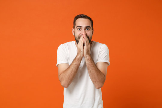 Surprised young man in casual white t-shirt posing isolated on bright orange wall background, studio portrait. People sincere emotions lifestyle concept. Mock up copy space. Covering mouth with hands.