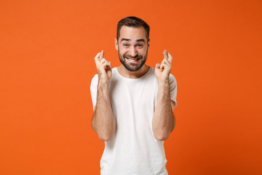 Excited young man in casual white t-shirt posing isolated on orange background in studio. People lifestyle concept. Mock up copy space. Waiting for special moment keeping fingers crossed, making wish.