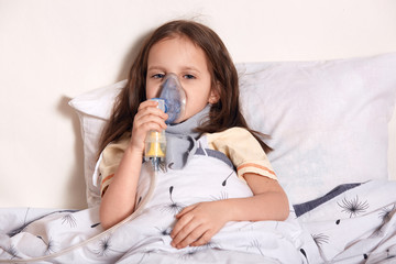 Closeup portrait of miserable poor child lying in bed alone, holding special equipment in one hand,...