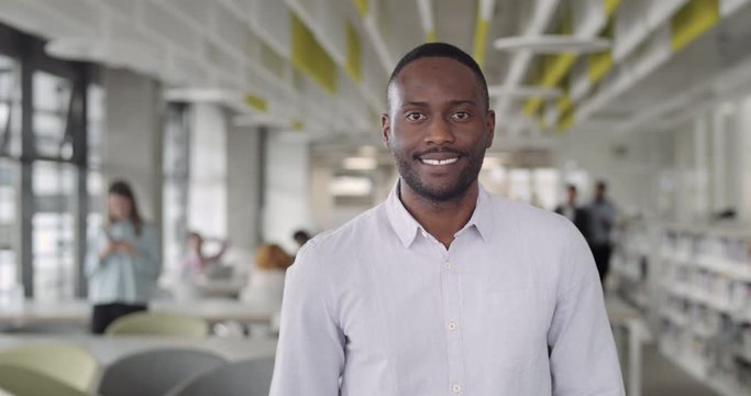 African male boss in shirt turning head to camera and smiling. Man in 30s team leader having good mood and standing in modern office working zone. Portraits. Blurred background.
