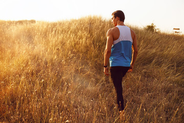 Naklejka na ściany i meble Image of a strong, athlete, muscular, fit, sexy runner man stands back in sportswear outside, isolated on a beautiful sunset background. Copy space.