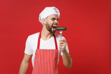 Crazy young bearded male chef cook or baker man in striped apron toque chefs hat posing isolated on red background in studio. Cooking food concept. Mock up copy space. Biting fresh cucumber on fork.