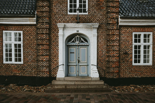 The Front View Of A Old Brick House With White Windows And Entrance With Light Blue Doors In The Oldest Denmark Town Ribe During The Autumn Rainy Day