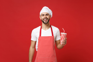 Cheerful young bearded male chef cook or baker man in striped apron white t-shirt toque chefs hat isolated on red background. Cooking food concept. Mock up copy space. Holding cup of cola or soda.