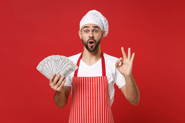 Shocked young bearded male chef cook or baker man in striped apron toque chefs hat isolated on red...