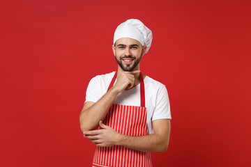 Smiling young bearded male chef cook or baker man in striped apron white t-shirt toque chefs hat posing isolated on red background. Cooking food concept. Mock up copy space. Put hand prop up on chin.