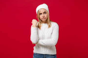 Aggression young woman in white sweater, hat isolated on red wall background studio portrait. Healthy fashion lifestyle people sincere emotions cold season concept. Mock up copy space. Clenching fist.