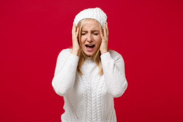 Frustrated young woman in white sweater, hat isolated on red background, studio portrait. Healthy fashion lifestyle, cold season concept. Mock up copy space. Keeping eyes closed, put hands on face.