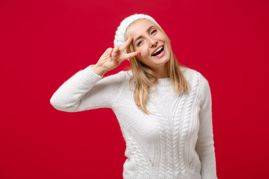 Cheerful young woman in white sweater, hat isolated on red wall background, studio portrait. Healthy fashion lifestyle, people emotions, cold season concept. Mock up copy space. Showing victory sign.