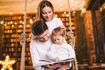 Obraz na płótnie Canvas Family togetherness. Mom, dad and son reading story book together sitting on swing evening home. Family and Parenthood Concept. Happy family reads childrens book while sitting home in loft interior