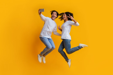 Fototapeta na wymiar Millennial Interracial Couple Jumping And Taking Selfiem Posing Over Yellow Background