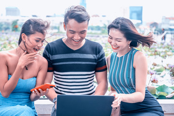 Smiling in front of a laptop screen. Three asian friends having happy outdoor time reviewing images on the computer