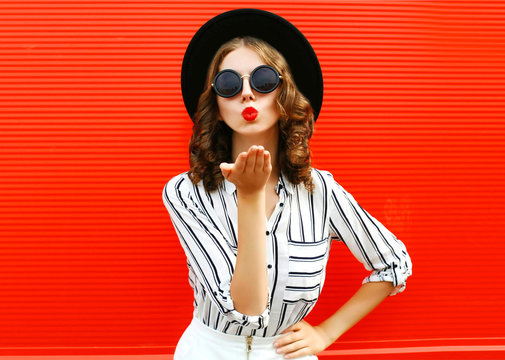 Portrait beautiful young woman blowing red lips sending sweet air kiss wearing white striped shirt, black round hat over red wall background