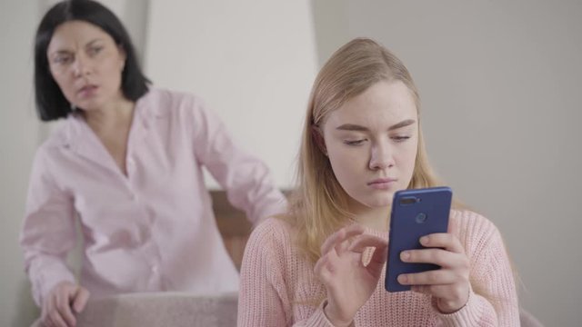 Camera approaching to worried Caucasian girl typing on smartphone as her mother looking at screen from the background. Shocked mom spying teenage daughter. Reliance, relationship.