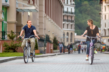 Happy smiling Caucasian family of father, mother and daughter have outdoor bicycle ride at summer on hotel buildings background. Active leisure and sport activities at resort.
