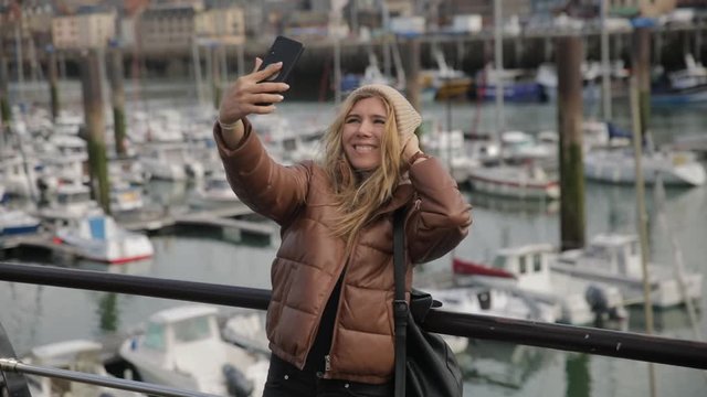 Happy young attractive stylish girl young woman blogger in port do a selfie at her cell phone with boats on background. Slow motion