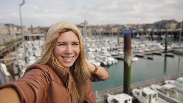 Happy Caucasian girl making selfie or a video call on a large lake, smiling and laughing into camera. Smartphone camera POV. Boats of port on background