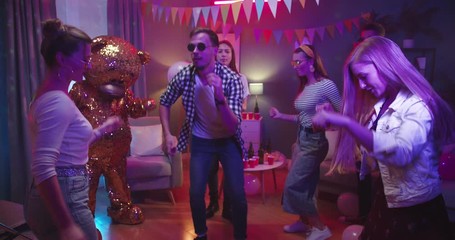 Joyful young Caucasian guy dancing and moving while having fun at the home party among best friends and golden grown teddy bear doll.