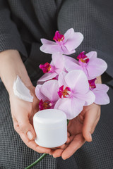 Background with blank white cream bottle and branch of beautiful pink orchids in a female hands. Branding mock-up. Vertical photo.