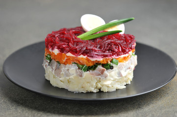 A classic salad of Russian cuisine - a herring under a fur coat. A portion of salad is decorated with green onions and a quail egg. Close-up. Black plate. Gray background.