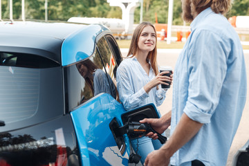 Traveling. Young couple traveling by electric car stopping at charging station boyfriend plugging...