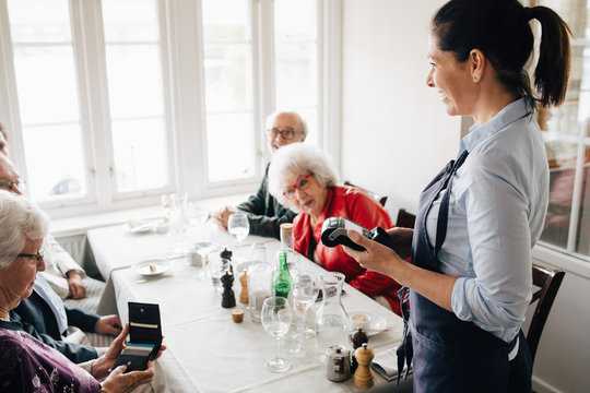 Happy owner standing with credit card reader by table while senior friends sitting in restaurant