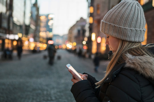 Teenage girl wearing knit hat text messaging on smart phone in city during sunset