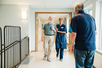 Senior man walking arm in arm with female nurse while looking at friend standing in alley at nursing home