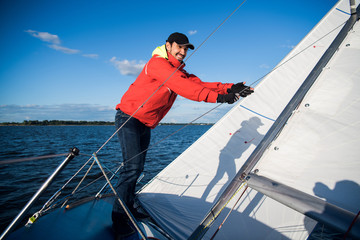 Young sailor in sunglasses and cap holds and moves ropes with both hands. He is calm and concentrated. Young man is preparing yacht for sailing. It is sunny outside.