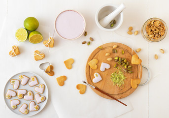 Iced spicy shortbread cookies in heart shapes, embellished with lime-raspberry icing and crushed pistachio nuts. Baking with love for Valentine day or advent and Christmas. White wooden table.