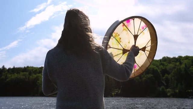 A backlit view of a woman with long hair blowing in the wind, seen from behind, playing an indigenous powwow drum, seeking enlightenment in nature