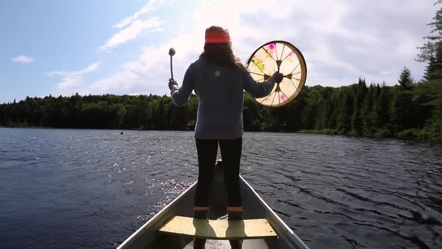 A backlit view of a woman standing on a canoe over a calm lake with lens flare, playing a sacred drum, worshipping nature with copy space to sides