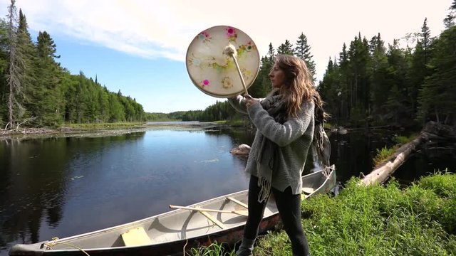 A spiritual lady is seen standing from the side, seeking divination and enlightenment in nature, playing a sacred native drum and beater by a lake