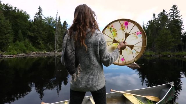 Slow motion footage from behind a spiritual woman seeking harmony and mindfulness in nature, standing on a canoe and playing a sacred native drum