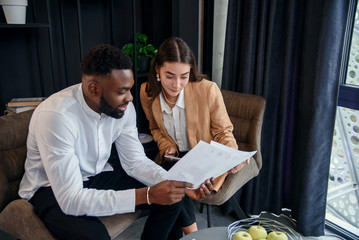 Business couple of african american man and european woman which sitting in specially designated room and using paper documents and tablet pc, conversing about their business project and their