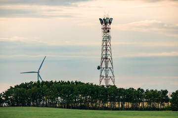 A wide angle late afternoon view of a cell site tower in a lush green landscape with woodland,...