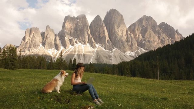young women hipster freelancer with dog border collie typing on computer keyboard, moving camera, working on laptop pc in outdoors Italy dolomites