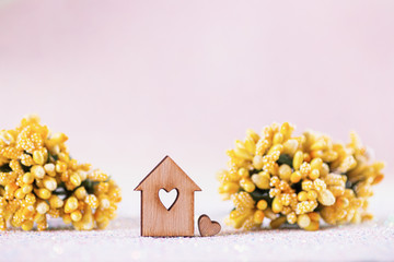 Closeup wooden house with hole in form of heart surrounded by spring yellow flowers on light pink background.