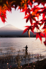 Silhouette of man fishing in lake with backdrop of Mount Fuji. View through red Autumn leafs