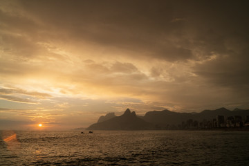 Naklejka na ściany i meble A dramatic sunset over Ipanema Beach in Rio de Janeiro, with golden light shining on grey clouds.