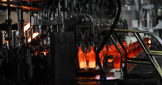 Manufacturing with automated conveyor robotic line for production of glass bottles on industrial factory. Hot glowing glass bottles on conveyor in machine moving passing by fire. Equipment in plant.