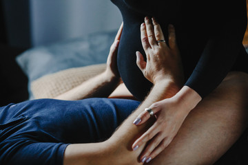 Pregnant woman sits on her husband, and he holds hands on her belly