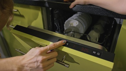 Mother teaching daughter smart girl learning to use dishwasher. Stylish modern Built In Kitchen Appliances in green black. Young mistress children loading putting dirty dishes in automatic dishwasher.