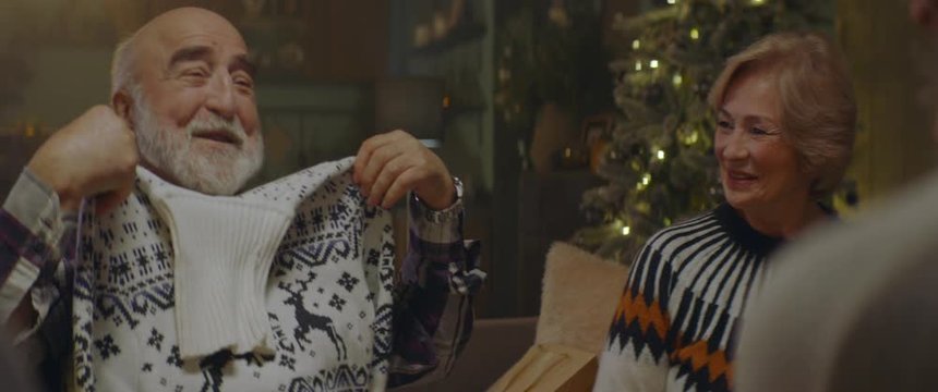 Senior Adult Caucasian Couple Celebrating Christmas Together With Their Family, Exchanging Gifts At Home, Old Man Receiving A Sweater From His Wife. Shot On ARRI Alexa Mini With Cooke 2x Anamorphic Le