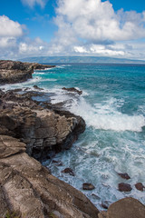 Waves crash on the rocks of a jagged coastline on a tropical island with clear blue water and a distant island visible.