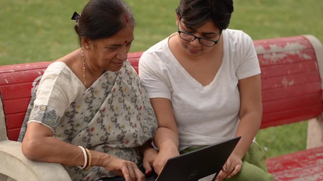 Young Indian woman helping her old Indian Bengali mother on a laptop sitting on a red bench in a park in Delhi Concept shot digital India and technology literacy for senior citizens