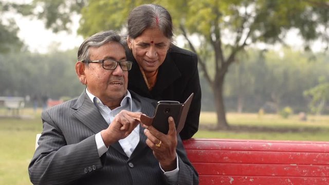 Happy retired senior citizen couple sitting outdoor in a park. Wife reading / working on a mobile, reading jokes and talking with her husband, smiling, laughing in a park in New Delhi, India